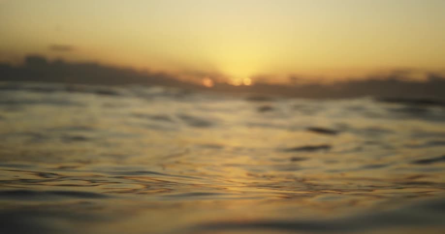 The camera floats at water level capturing ripples and gentle swells of the ocean surface as the sun sets behind a hazy distant coastline casting a warm golden glow across the moving water and the soft clouds in the horizon sky