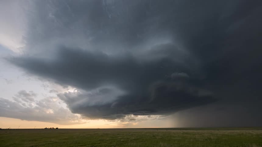 A massive dark supercell storm cloud looms over a flat grassy plain at sunset