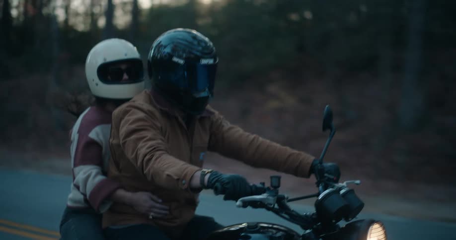 A man and woman ride a black motorcycle along a road at dusk both wearing protective helmets the woman sits behind the man holding onto his waist as they travel past a wooded area