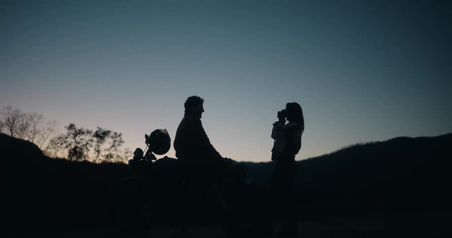 A couple stands outdoors beside a motorcycle at evening talking after sharing a hug