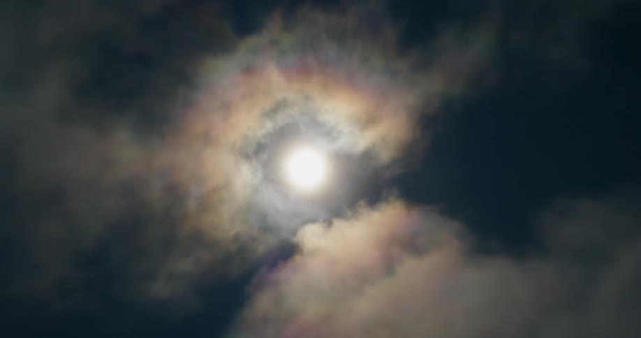 The sun burns through dark swirling clouds creating a luminous halo with subtle rainbowlike diffraction that gives the sky an otherworldly atmospheric glow
