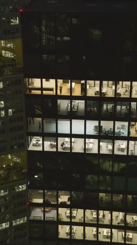 A person walking seen through the window of a skyscraper at night with most of the office lights glowing inside the building