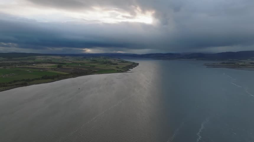 Coastal terrain with green fields on the left and calm waters on the right under dramatic cloudy sky