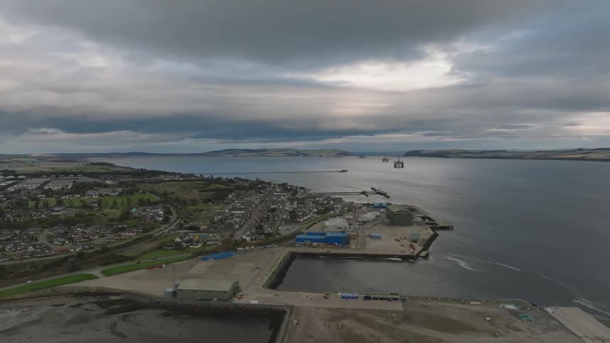 A highangle aerial shot shows a coastal town and industrial harbor under a cloudy sky featuring residential blocks port buildings and large structures in the water near the horizon