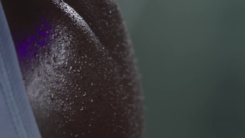 A persons muscular shoulder glistening with sweat where fine droplets catch the light against dark skin and soft fabric at the edge of the frame conveying heat effort and physical intensity