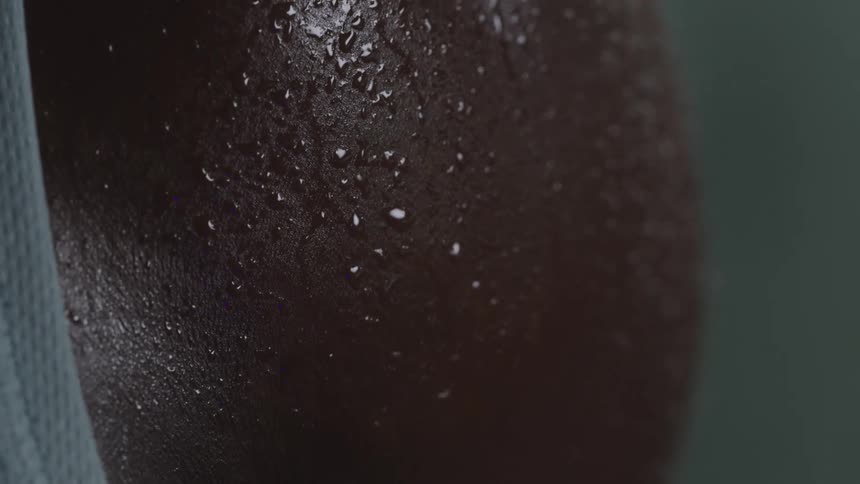 A human shoulder and upper arm reveals fine body hair and tiny beads of moisture catching soft moody light with subtle purple highlights accentuating skin texture against a dark outoffocus background