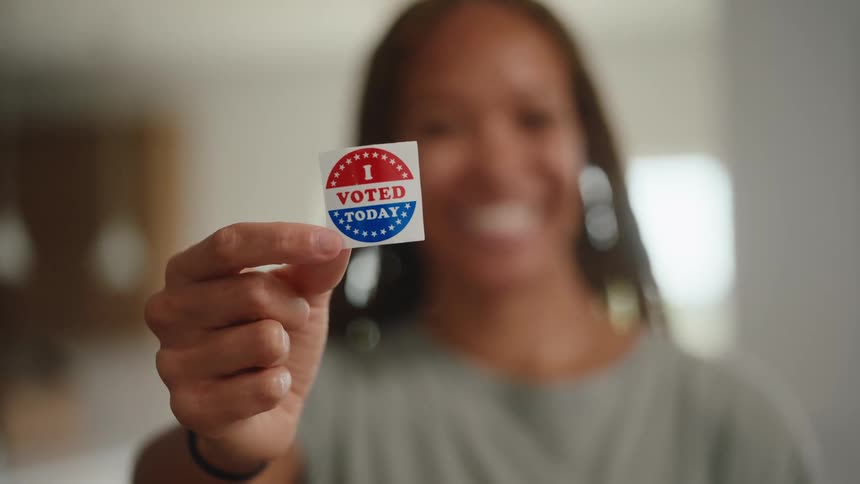 A smiling woman holds up a red white and blue i voted today sticker toward the camera in a brightly lit indoor room
