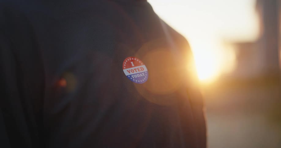 A person stands outdoors at sunset wearing a dark shirt with an i voted today sticker pinned to their chest the golden sunlight flares across the frame highlighting the texture of the fabric and the patriotic colors of the badge