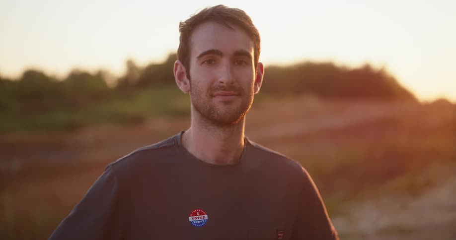 A young smiling man is standing outdoors in a rural area with dirt grounds and trees