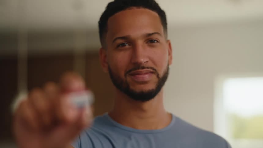 A softly smiling man stands indoors with his face out of focus while holding a small red white and blue object