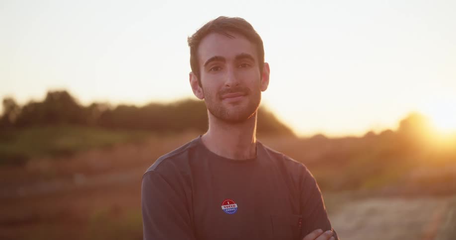 Man with his arms crossed stands in a field with the sun setting on the horizon