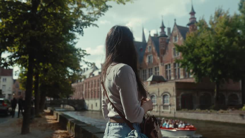 Woman seen from behind looks toward an urban canal with historic buildings in the background holding a bag near a treelined bank
