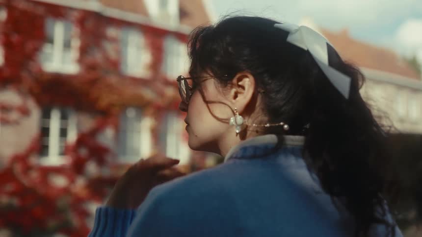 A woman with dark hair tied in a white ribbon walks along a canal in a historic city passing a red ivycovered house and a stone bridge under bright daylight