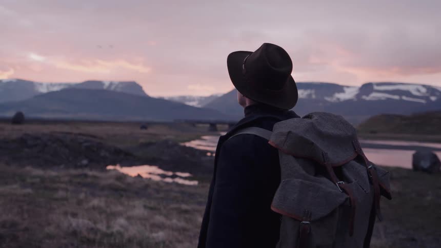 Person wearing a backpack and hat looks over a wide landscape with a winding river and mountains in the background at dusk