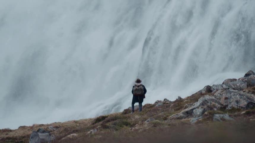 Man stands on a hill watching a waterfall flow down a mountain cliff