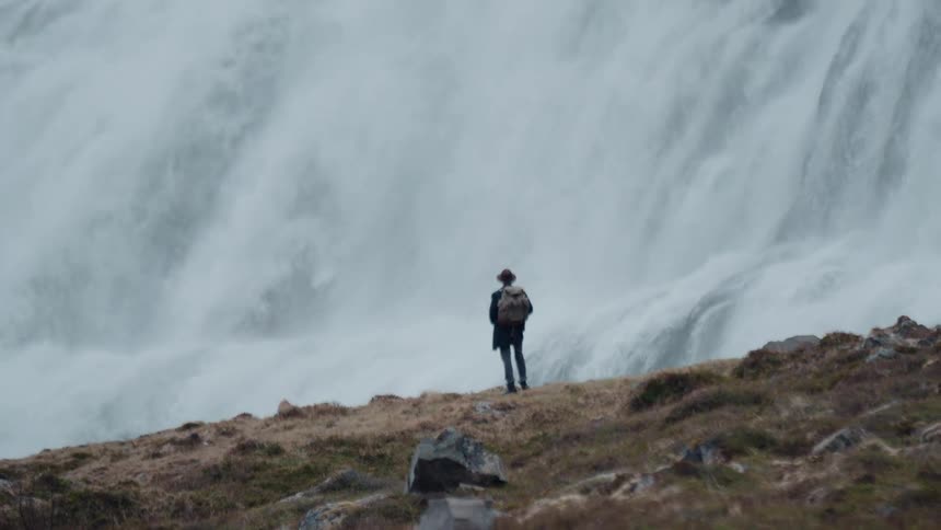 Man stands at the edge of a mountain watching a waterfall flow down a mountain cliff