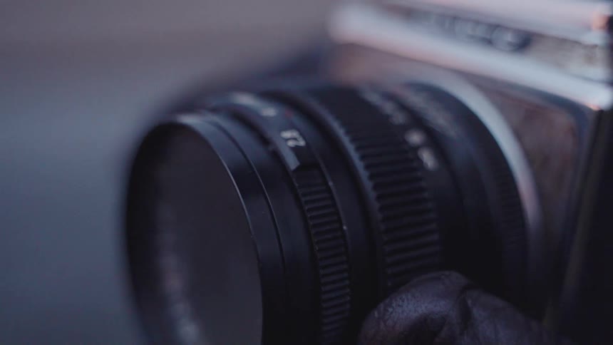 A closeup shot shows a hand in a black glove adjusting the focus and aperture rings of a vintage film camera lens the camera slowly pulls back to reveal more of the metallic body and textured grip as the settings are dialed in