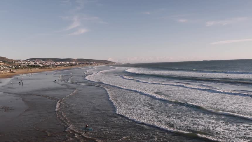 A long curving coastline unfolds beneath a clear sky as parallel lines of whitecapped waves roll steadily toward a broad sandy beach dotted with small figures and backed by a distant seaside town