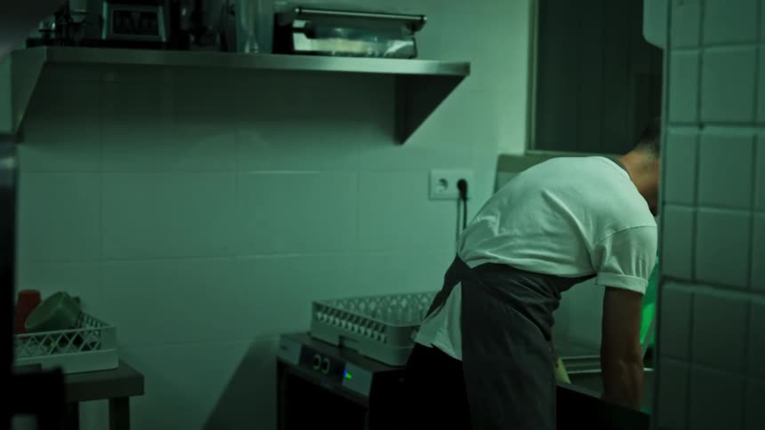 A man wearing a white tshirt and grey apron stands at a commercial sink in a dimly lit kitchen scrubbing dishes and moving items while green soap sits on the counter beside him