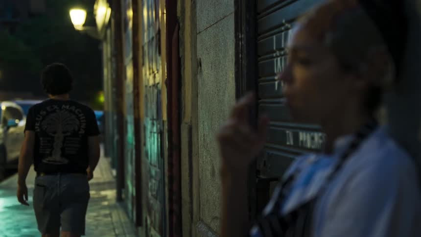 Woman standing outside of a building smoking as a man walks out of the door and lights his cigarette then walks down the sidewalk and the woman goes back inside