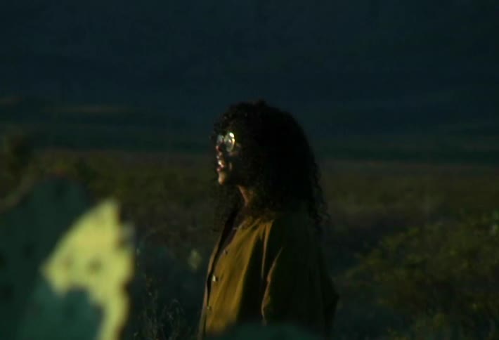 A man stands among desert plants talking to a black woman framed by distant mountains under a clear blue sky