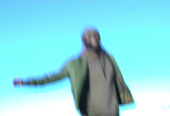 A joyful black man in a green jacket and beanie jumps repeatedly into the air on a field against a bright blue sky laughing and cheering
