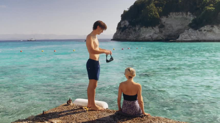 A young man stands on a rocky ledge putting on a snorkel mask while a blonde woman sits beside him both looking out over clear turquoise water toward a lush cliff and distant boats in a calm bay during a bright sunny day