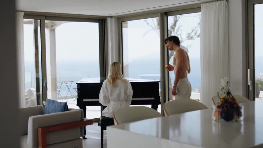 A woman sits at a piano in a bright seaside living room while a shirtless man stands nearby holding a piece of fruit with large windows opening to an ocean view