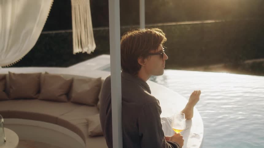 A person relaxes drinking wine by an infinity pool at golden hour holding a drink with one foot dipped in the water framed by soft cushions flowing curtains and warm sunlight