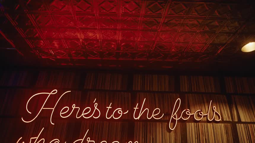 A neon sign displaying the phrase heres to the fools who dream glows in orange light against a wall of vertical vinyl records positioned beneath a decorative tin ceiling illuminated by a soft red ambiance