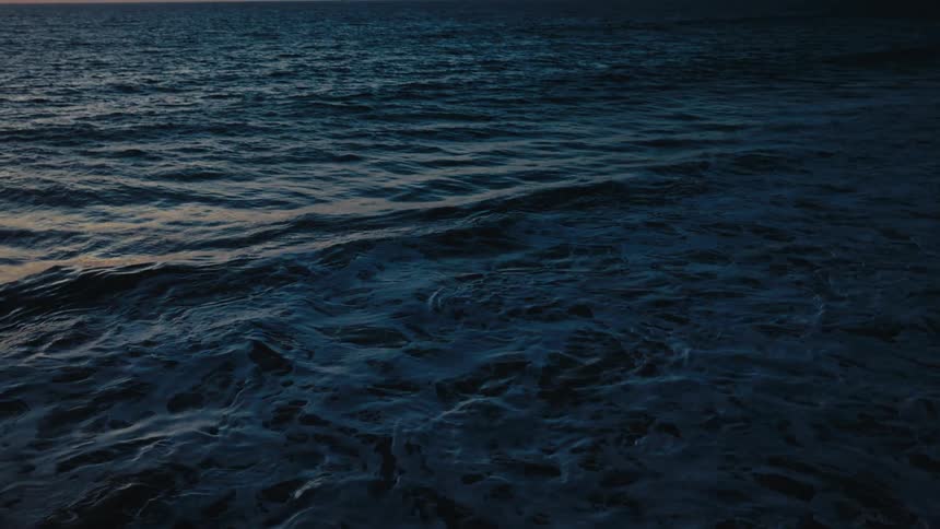 Dark ocean waves roll toward the camera during twilight while the orange glow of a sunset lingers on the horizon over distant mountains and the faint lights of anchored cargo ships