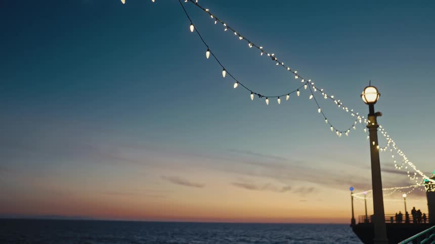 Several silhouetted people stand and stroll along the piers deck beneath glowing string lights quietly watching the ocean as dusk settles over the horizon