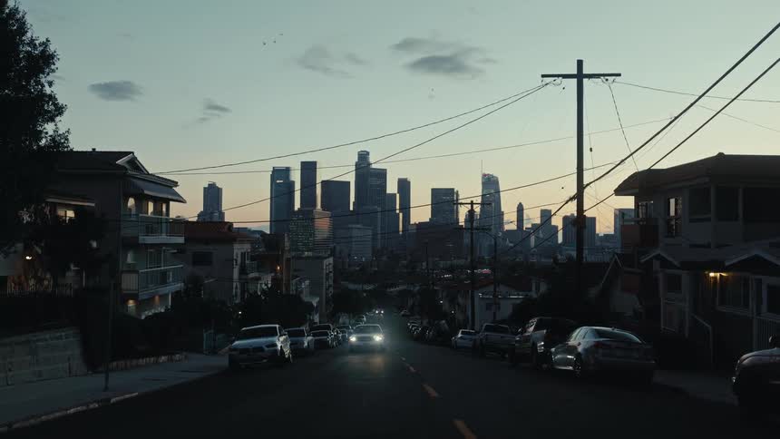 A car with its headlights on driving down a city street at evening time with parked cars lining both sides