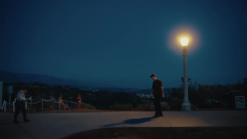 Man briefly jumps onto the base of a streetlamp overlooking an urban valley before passing a woman in uniform walking in the opposite direction at night
