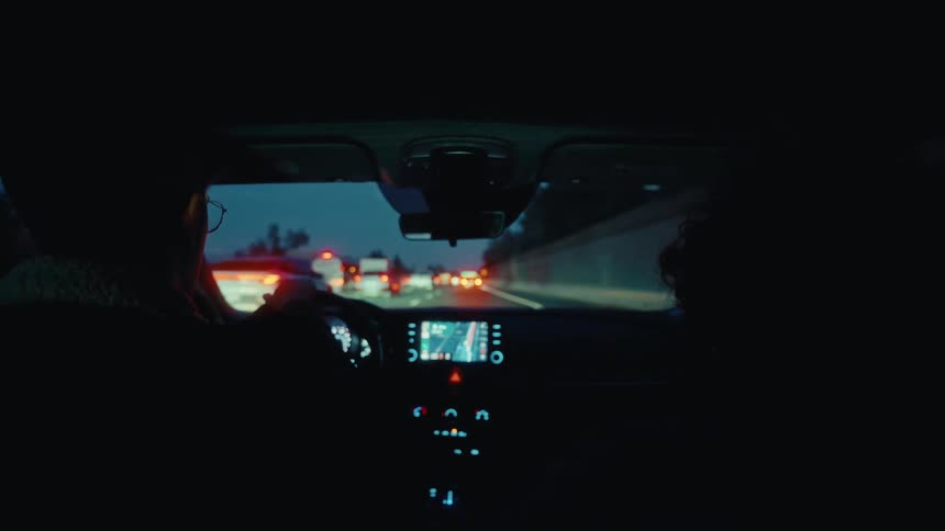 A man with glasses drives a car on a crowded highway at dusk while a passenger sits beside him showing the dashboard lights and tail lights of traffic through the windshield as they move along the freeway