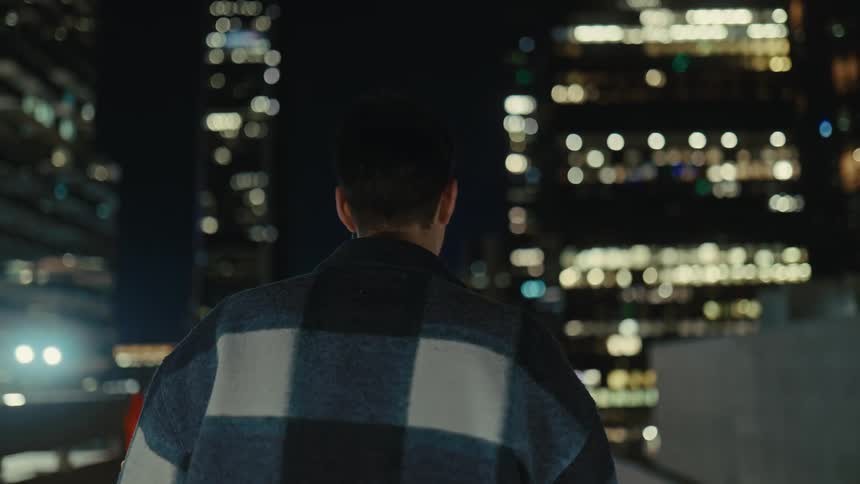 A person in a plaid jacket stands against a softly glowing city skyline at night then walks away as the lights blur into the distance behind them