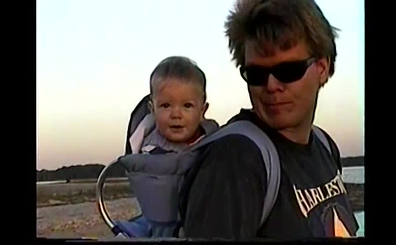 A man wearing sunglasses carries a smiling baby in a blue child carrier backpack while standing on a rocky shoreline near the water during sunset