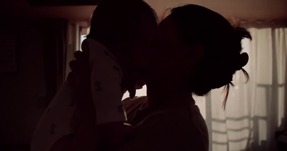 A mother holds her infant up toward a window gently nuzzling and kissing the babys face in silhouette as the warm evening light glows behind them in a domestic room