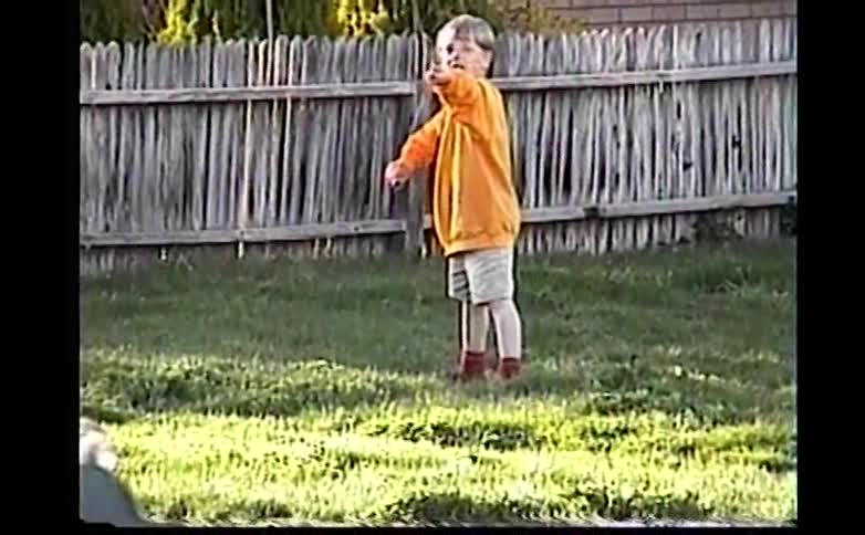 A young boy wearing an orange sweatshirt and tan shorts plays with a thin stick in a grassy backyard near a wooden fence during the day