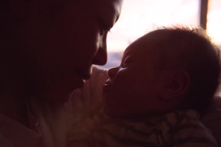 A mother gently cradles her newborn baby by a window during sunset smiling and touching her nose to the infants face while the city skyline remains visible in the softfocus background