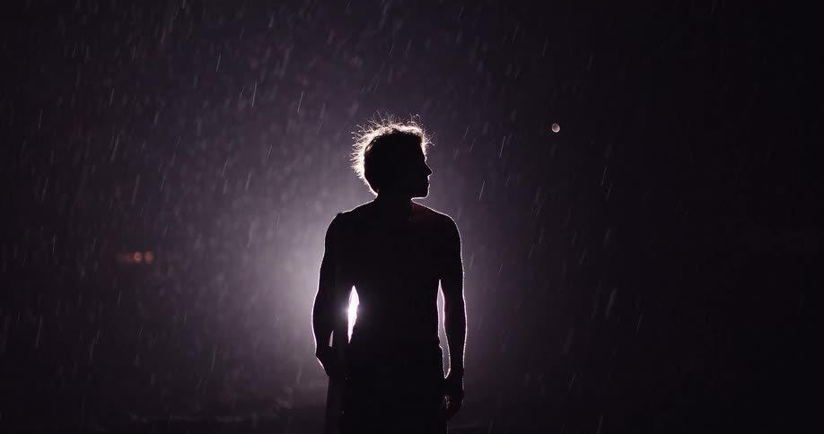 Backlighting casts a male surfer holding a surfboard and standing in a rainstorm at night into silhouette