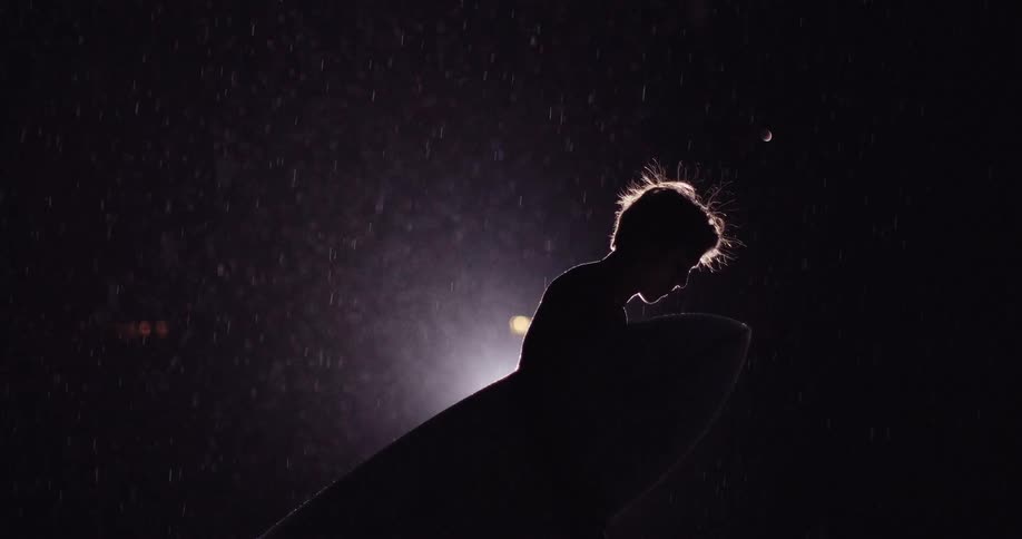 A silhouetted surfer stands in the rain at night holding a board against their body as backlight glows through falling droplets