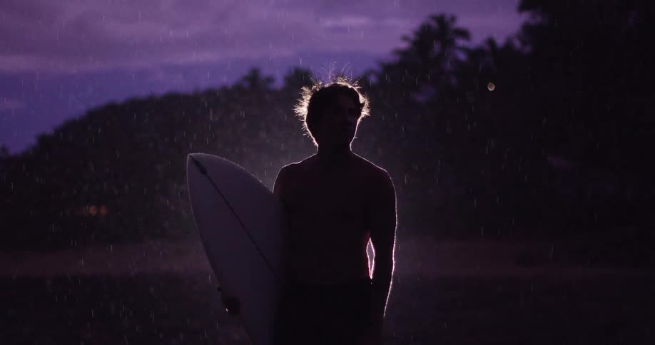 Young man standing along the coast at night holding his surfboard in the rain as lighting flashes illuminate the sky