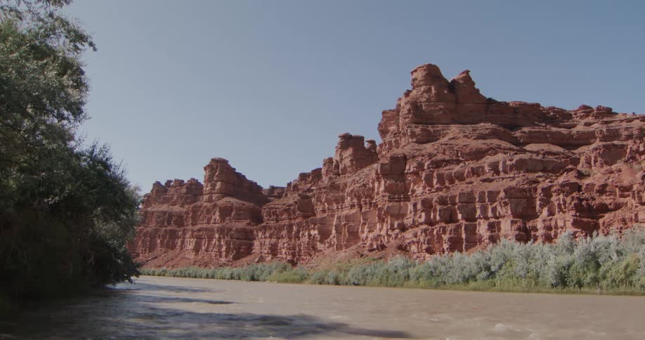 Tall red rock cliffs with jagged formations tower over a muddy river lined with green trees and bushes under a clear blue sky