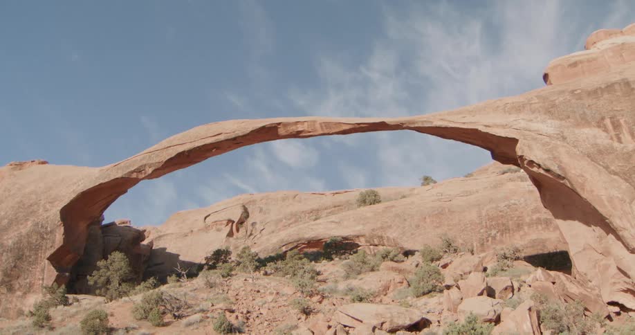 A landscape arch spans across a red sandstone canyon under a clear blue sky while sparse green shrubs and fallen rock debris sit at the base of the massive natural formation during a bright sunny day