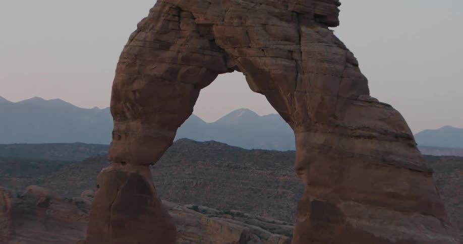 A large natural stone arch stands in a desert landscape during twilight with distant mountains visible through the opening under a clear sky