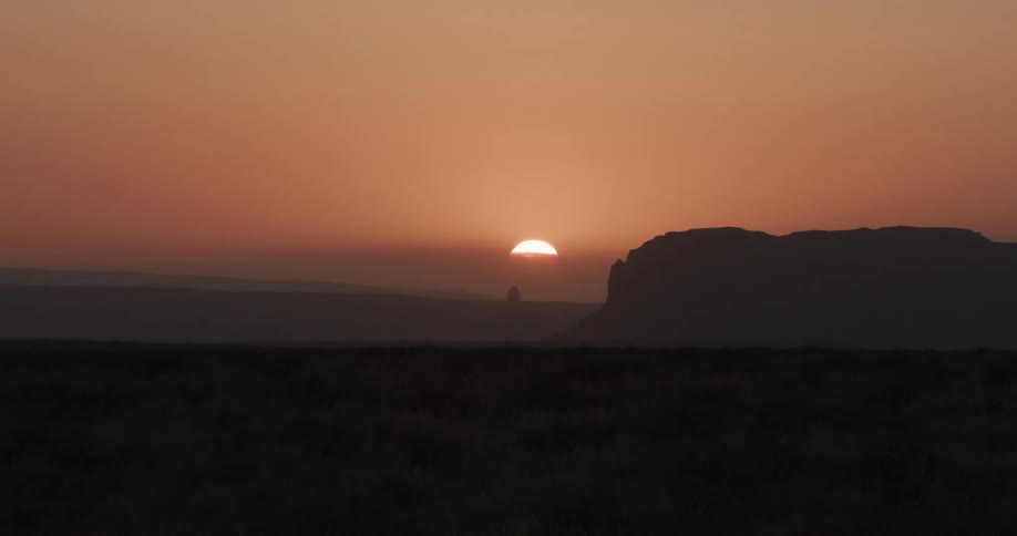 The sun sets slowly behind a dark mountain silhouette in a vast desert casting an orange glow across the hazy horizon and illuminating the dry brush in the foreground