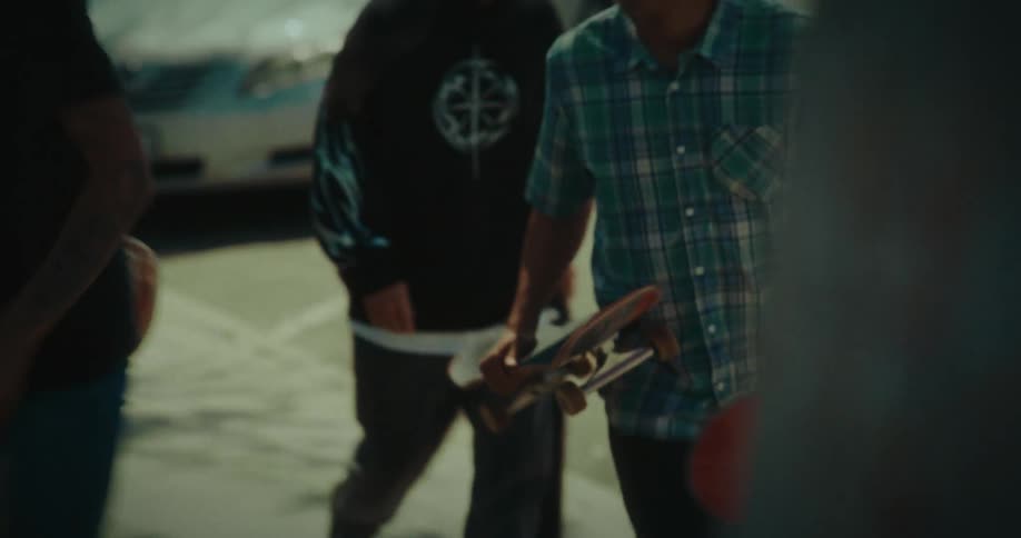 A group of young skaters carrying their boards walks along a sunny suburban sidewalk past parked cars talking and laughing together