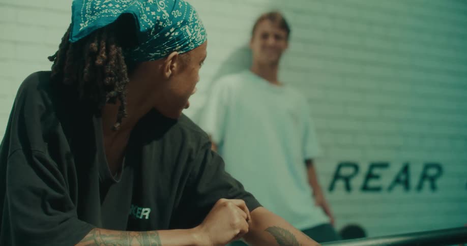 A woman with dreadlocks and a blue bandana talks to a man in a white tshirt leaning against a white brick wall outdoors