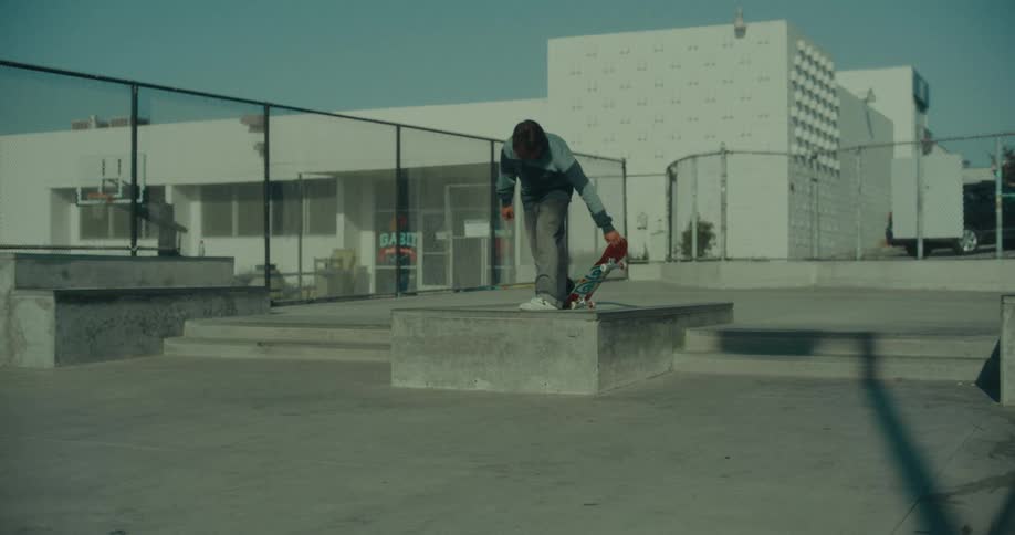 A man wearing a striped long sleeve shirt and grey pants jumps onto a concrete ledge with his skateboard at an outdoor skatepark near a white building with a basketball hoop